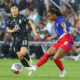 Jaedyn Shaw kicks a ball while wearing a blue and red national team kit. Ji So-yun runs behind her.