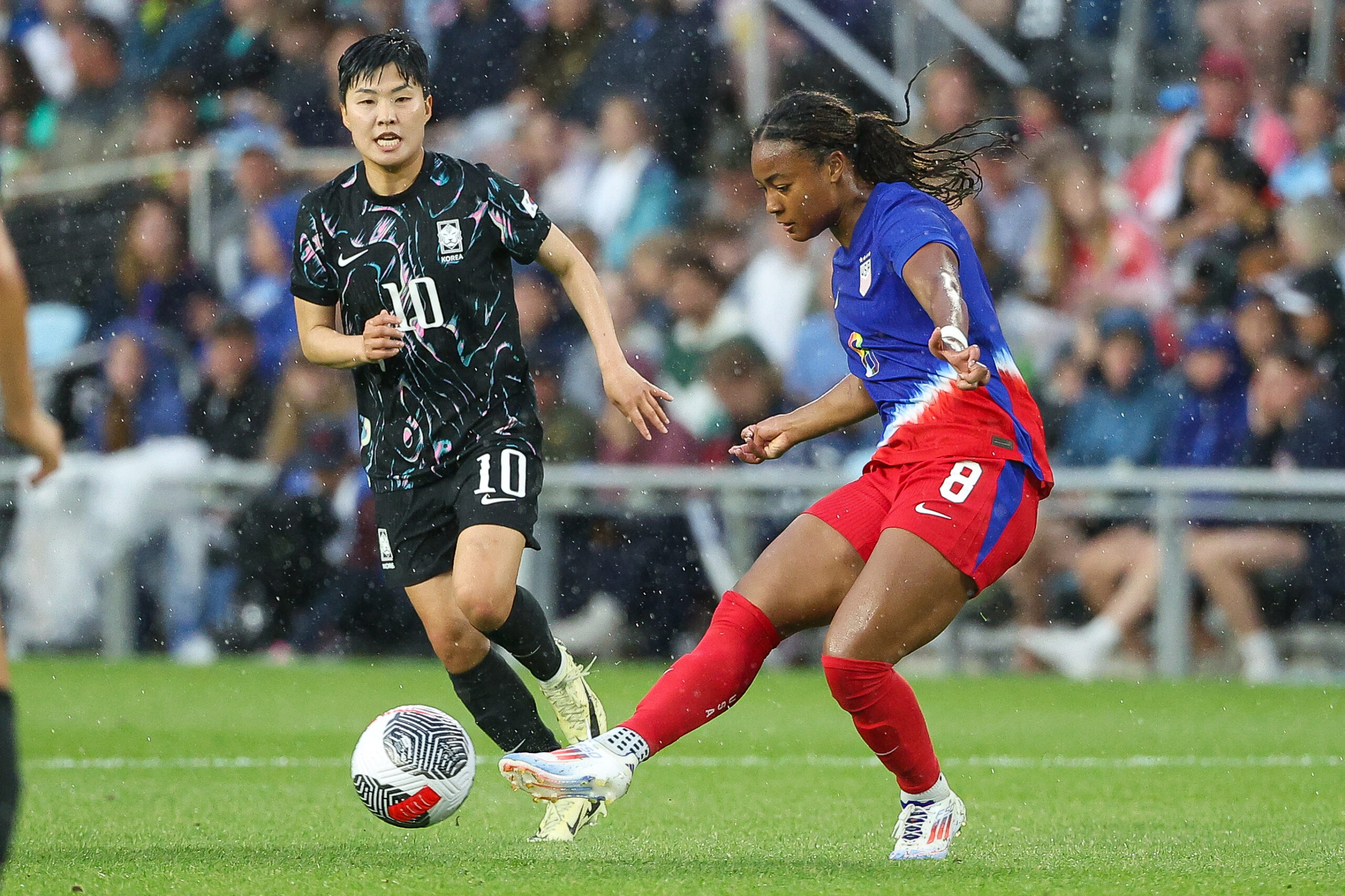 Jaedyn Shaw kicks a ball while wearing a blue and red national team kit. Ji So-yun runs behind her.